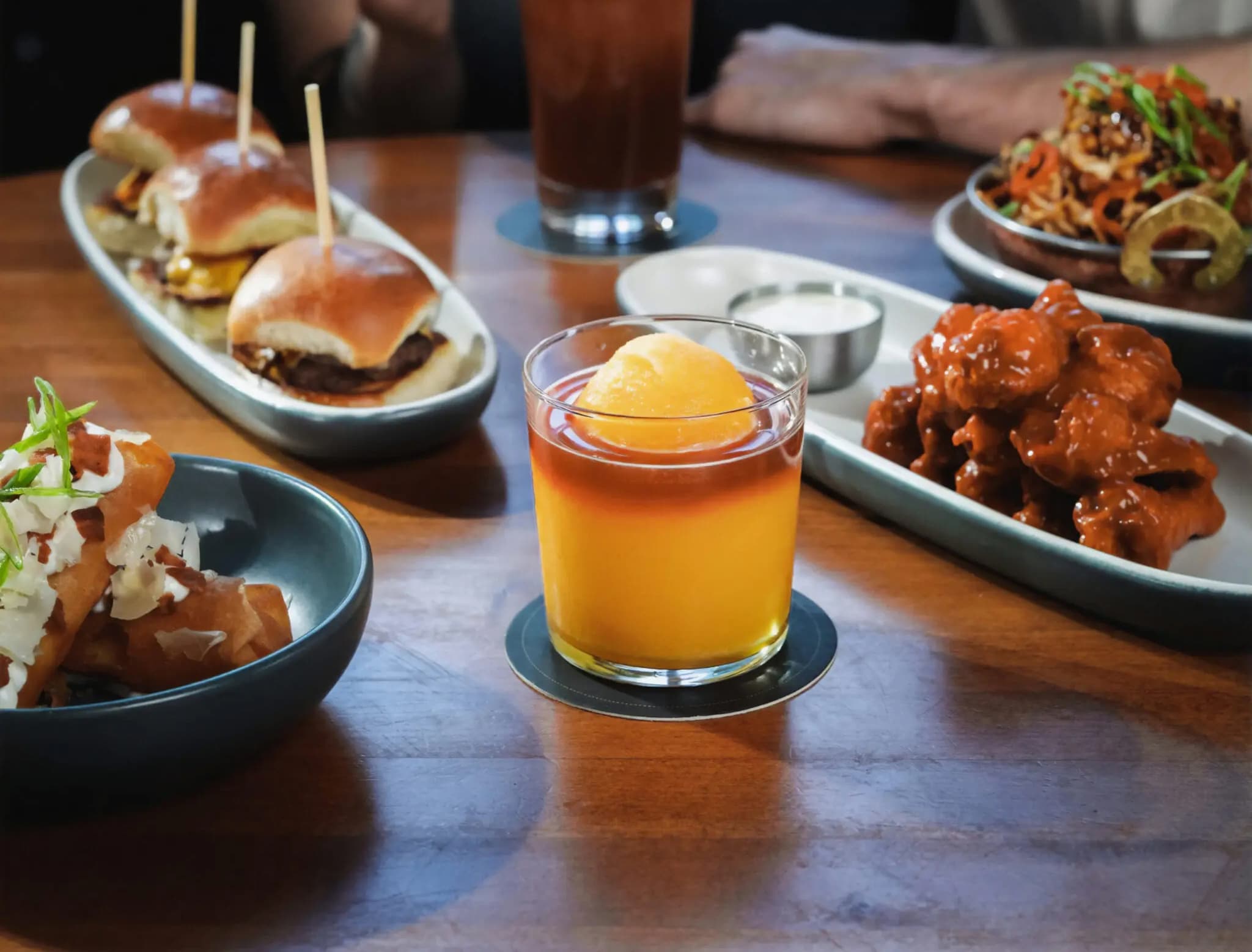 Real peach bellini slush cocktail surrounded by Hibachi chicken wings, burger sliders, crispy mashed potato spring rolls, and another dish in the background on a wooden table.
