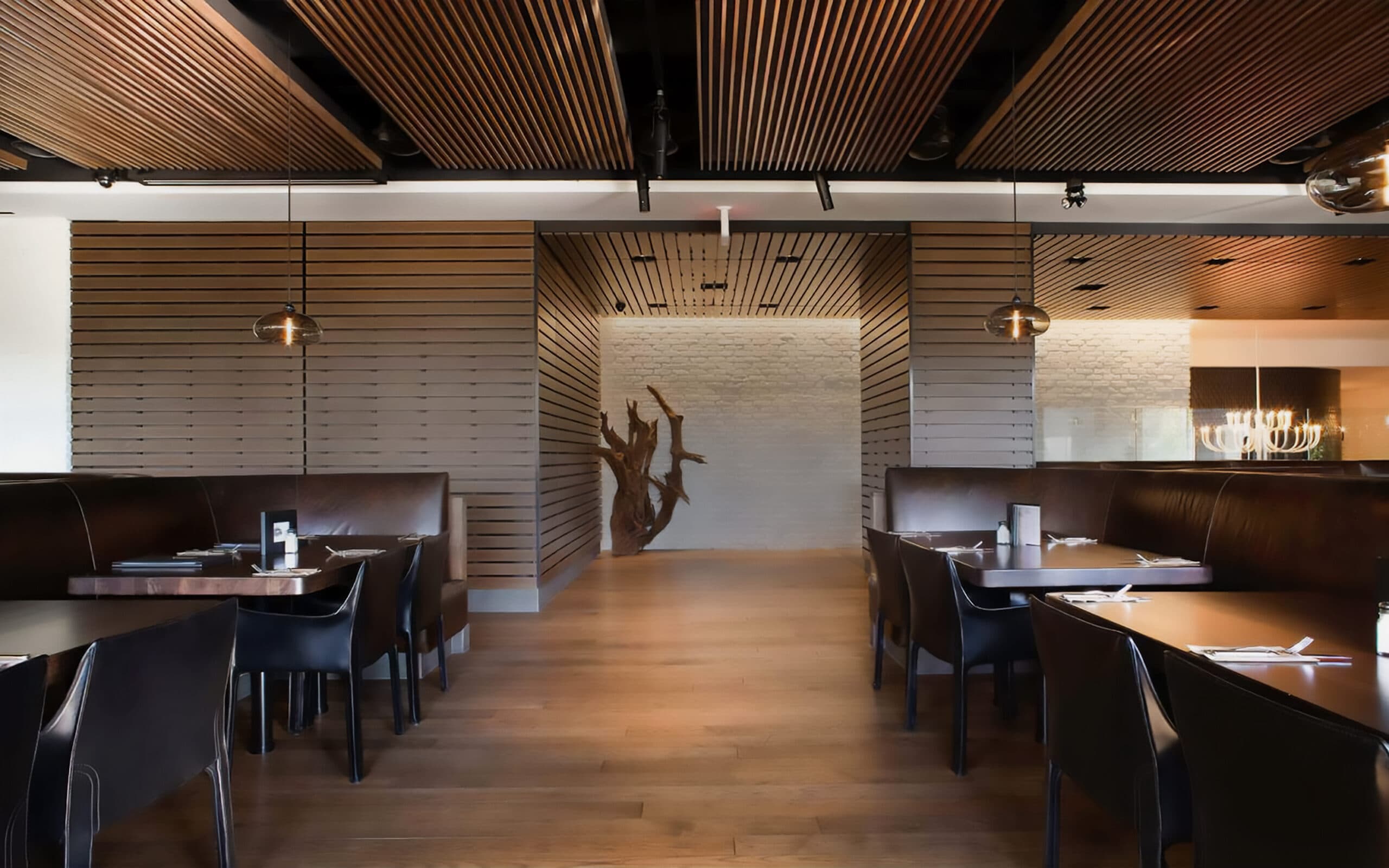 Modern restaurant dining area with leather booths, wooden tables, slatted wood ceiling and a tree sculpture display at the end of the central hallway.