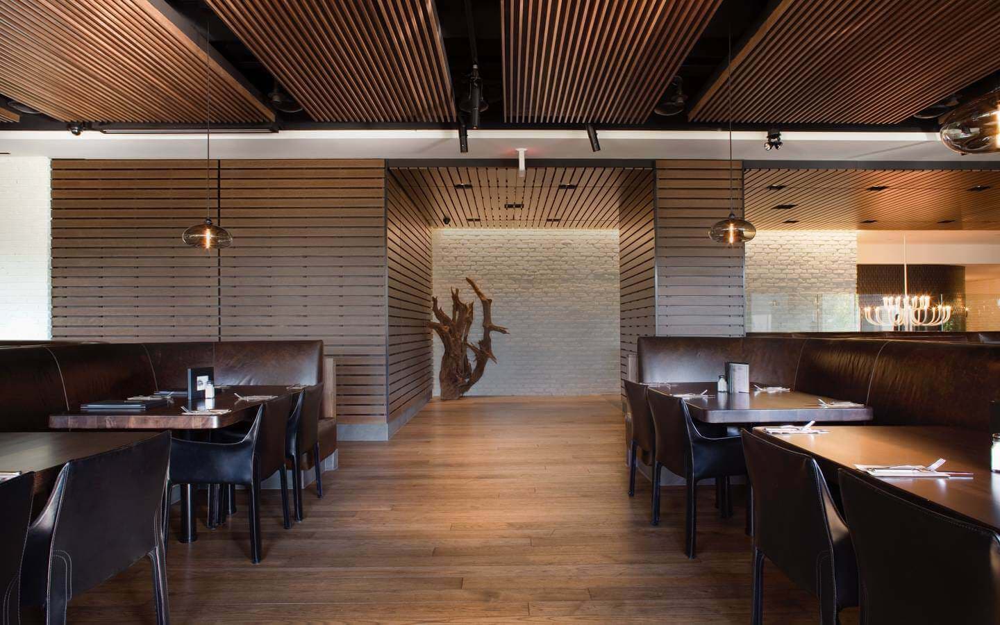 Modern restaurant dining area with leather booths, wooden tables, slatted wood ceiling and a tree sculpture display at the end of the central hallway.